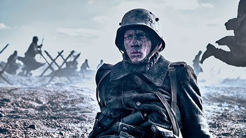 A dark image of a soldier sitting on the ground wearing uniform. His face is dirty. In the background is a war scene. Still taken from Netflix film All Quiet on the Western Front. Copyright - ReinerBajo - Netflix.