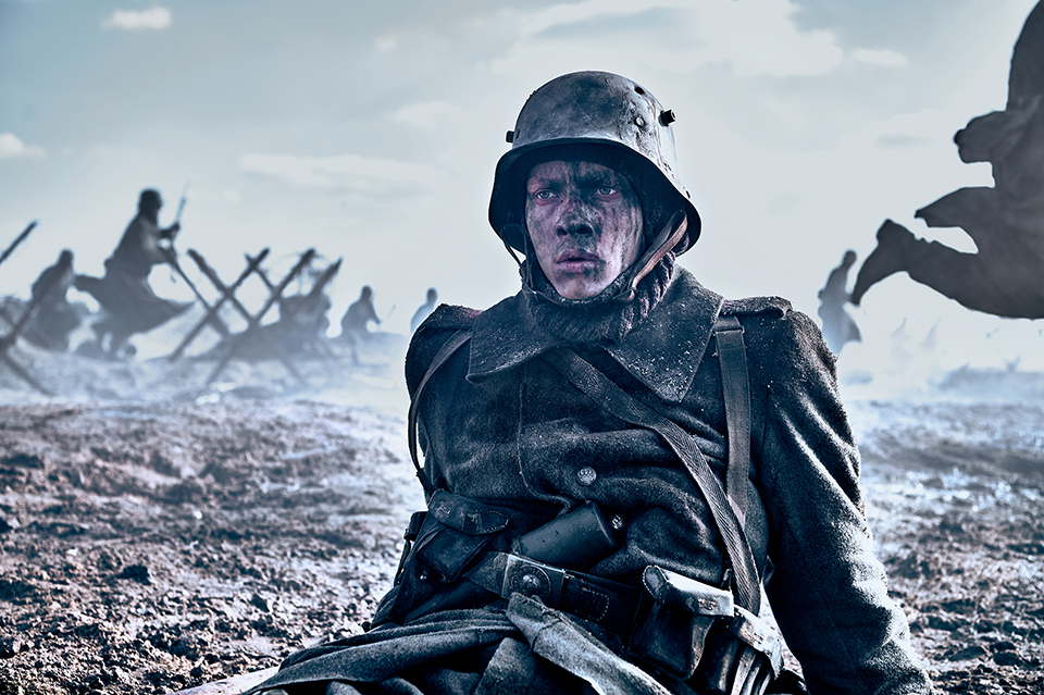 A dark image of a soldier sitting on the ground wearing uniform. His face is dirty. In the background is a war scene. Still taken from Netflix film All Quiet on the Western Front. Copyright - ReinerBajo - Netflix.