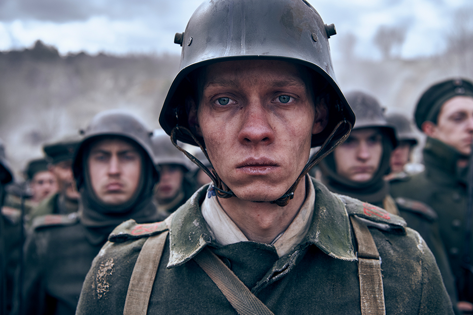 A soldier's face, head and shoulders. He looks into the distance. He is wearing uniform and a helmet. In the background there are other soldiers. Still taken from Netflix film All Quiet on the Western Front. Copyright - ReinerBajo - Netflix.