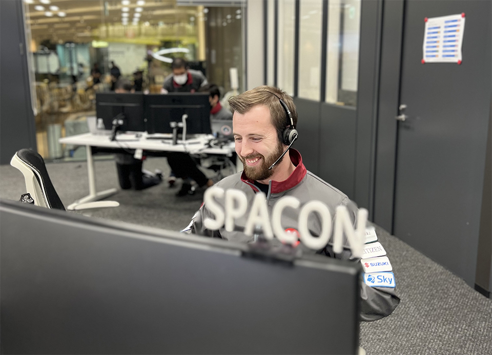 Federico Giusto is seated at a computer desk wearing a headset and smiling. On top of the computer screens is some lettering, which reads SPACON.A couple of other people are in the background at another desk with computers. 