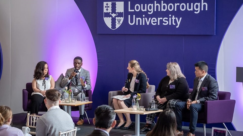 Five people are seated on a stage. Behind them is the University logo. One person is talking into a microphone as the others look on.