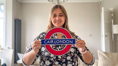 Tanya is smiling and holding up a London Underground logo sign but the text says 'CAIR.London'