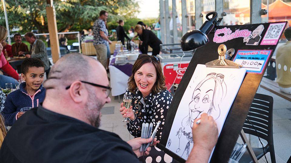 A person drawing a caricature of another person who is holding a champagne glass and smiling.