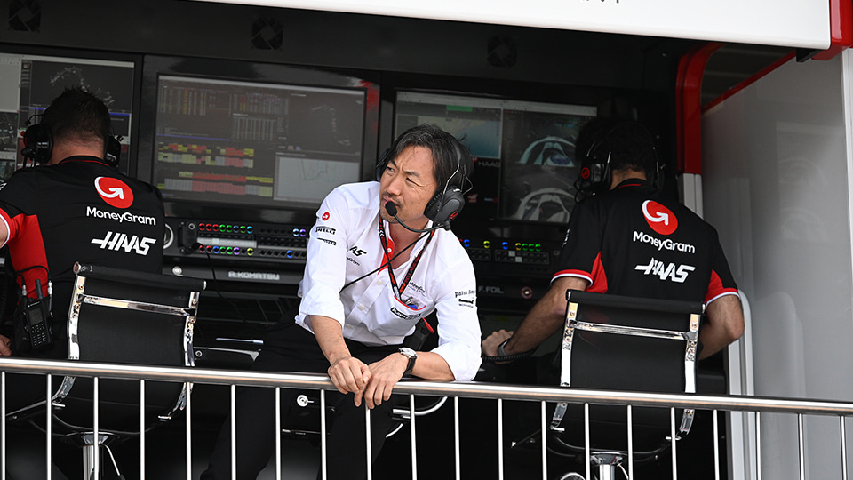 Ayao leaning over a railing wearing a headset. There are screens behind him people facing towards the screens on either side of him.
