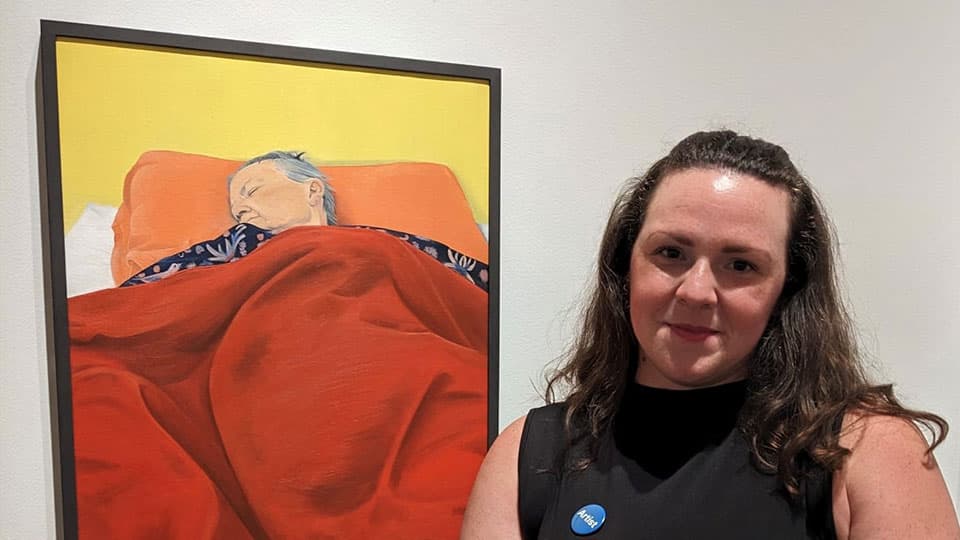 Laura is at The National Portrait Gallery in London standing in front of her painting called ‘I’ll Never Not Miss You’. The painting is in a black frame and the painting is of her mum asleep in bed with a red blanket over her.