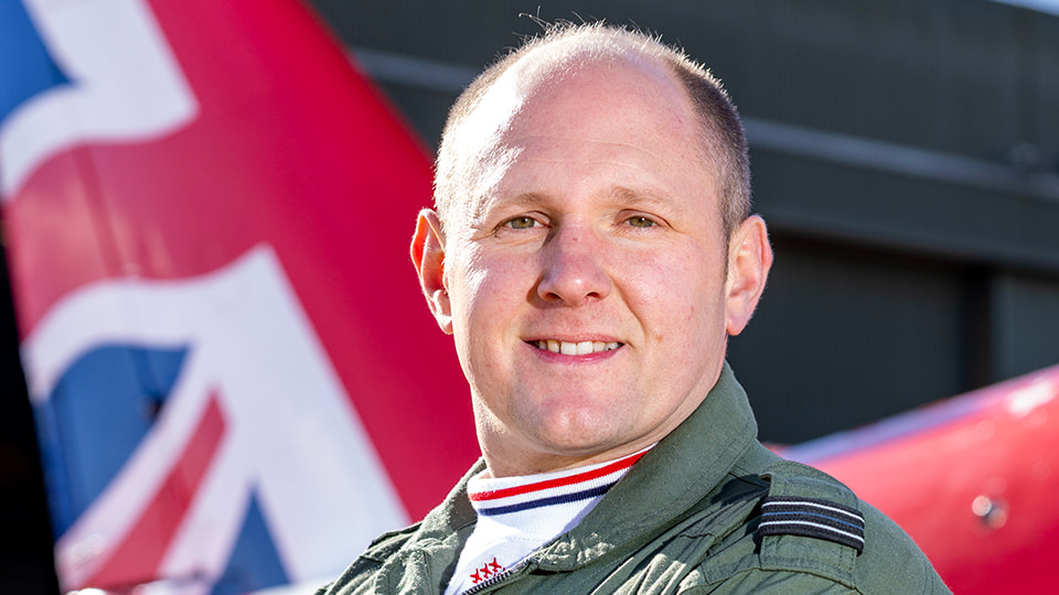 Flt Lt Rob Thomas wearing his green RAF Aerobatic Team suit. He stands in front of a red Arrows plane with his arms folded.