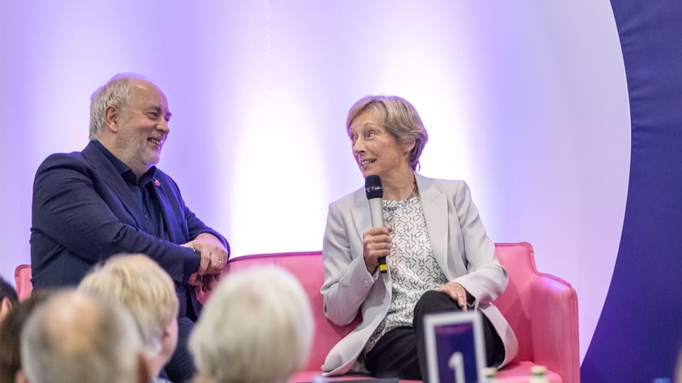 Professor Nick Jennings and Dame Liz Nicholl seated on a pink sofa. Liz holds a microphone and they are smiling.