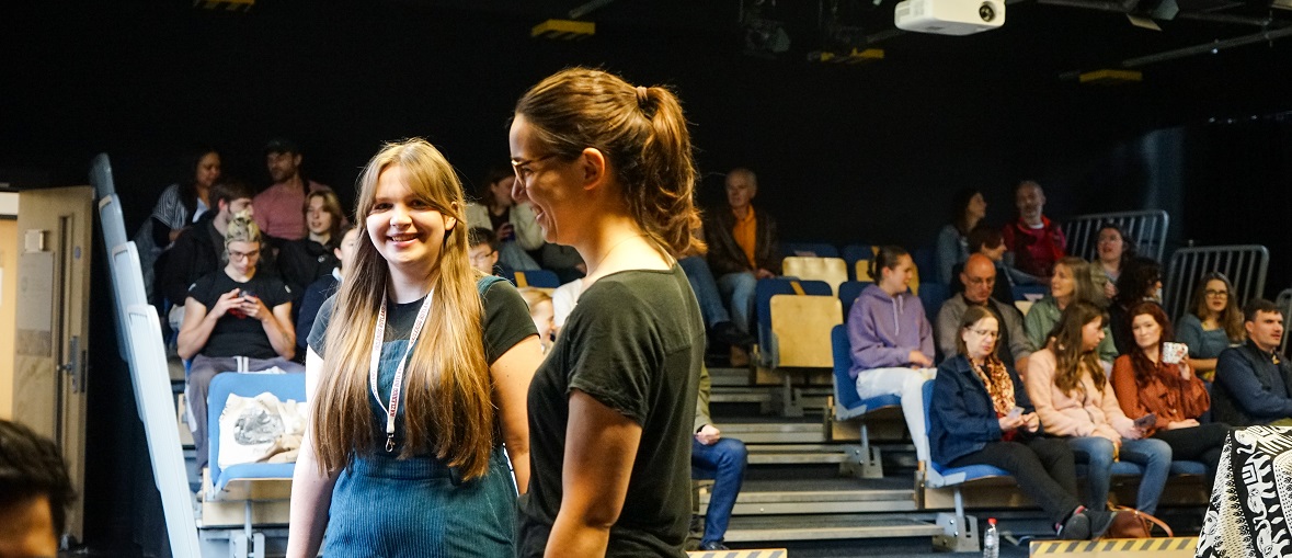 Two female students stand smiling in a theatre space, behind them a crowd of people are seated on bleacher seating