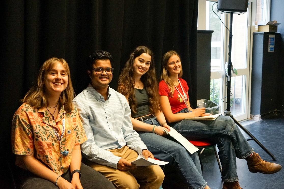 four students smiling sitting on chairs in a black studio space