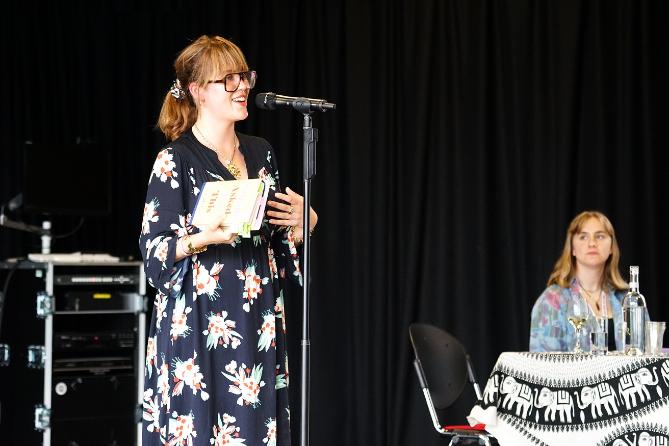 Charly Cox, a white woman in her 30s with brown hair tied in a ponytail wearing round glasses and a blue flowery dress, stands and talks into a microphone, a student sits to the right of her on a table