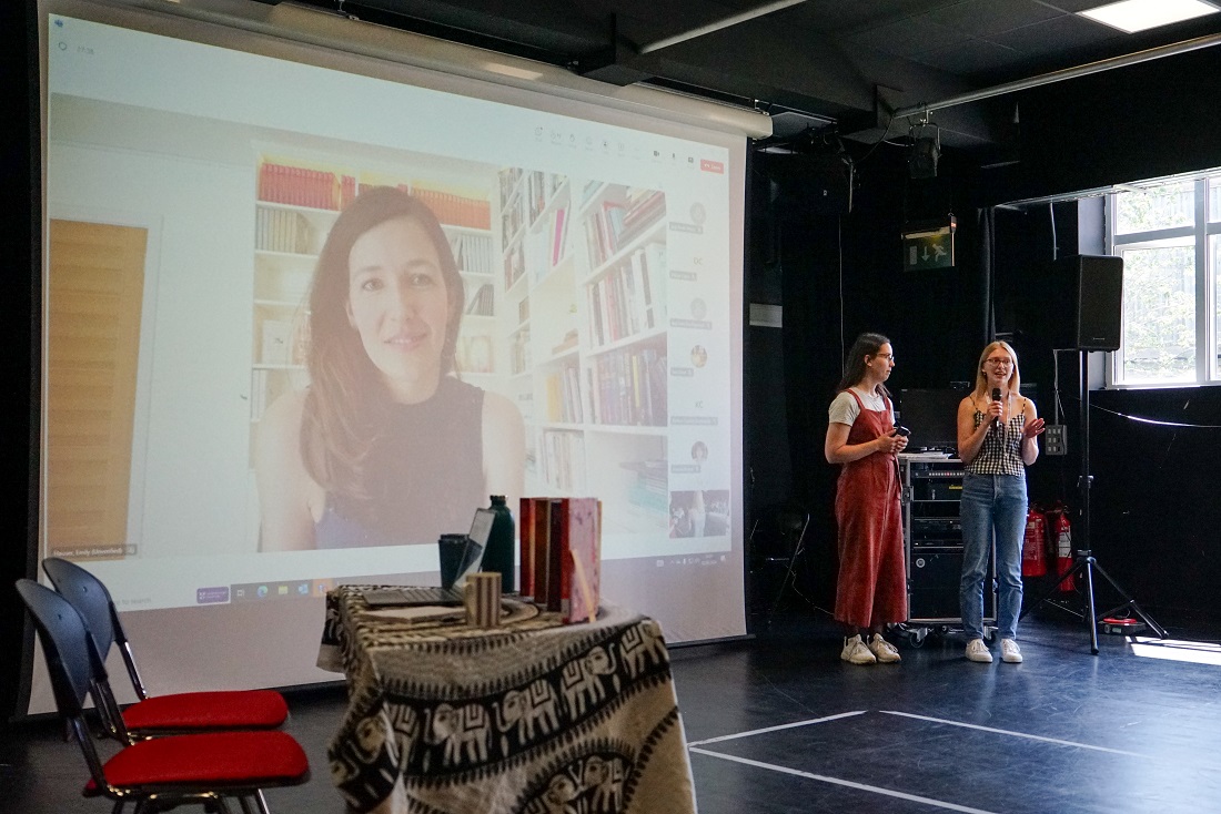 Emily Hauser, a white woman in her 30s with brown hair, appears on a large projector screen, to her right, two students stand and talk into a microphone