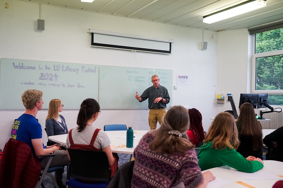 A white walled teaching room with a whiteboard. A man with grey hair gestures to the board to a group of students sitting around tables in front of him.