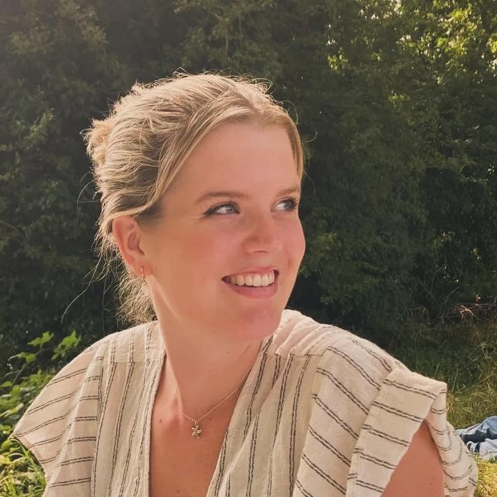 A student with blonde hair tied up smiling wearing a beige top with bushes in the background
