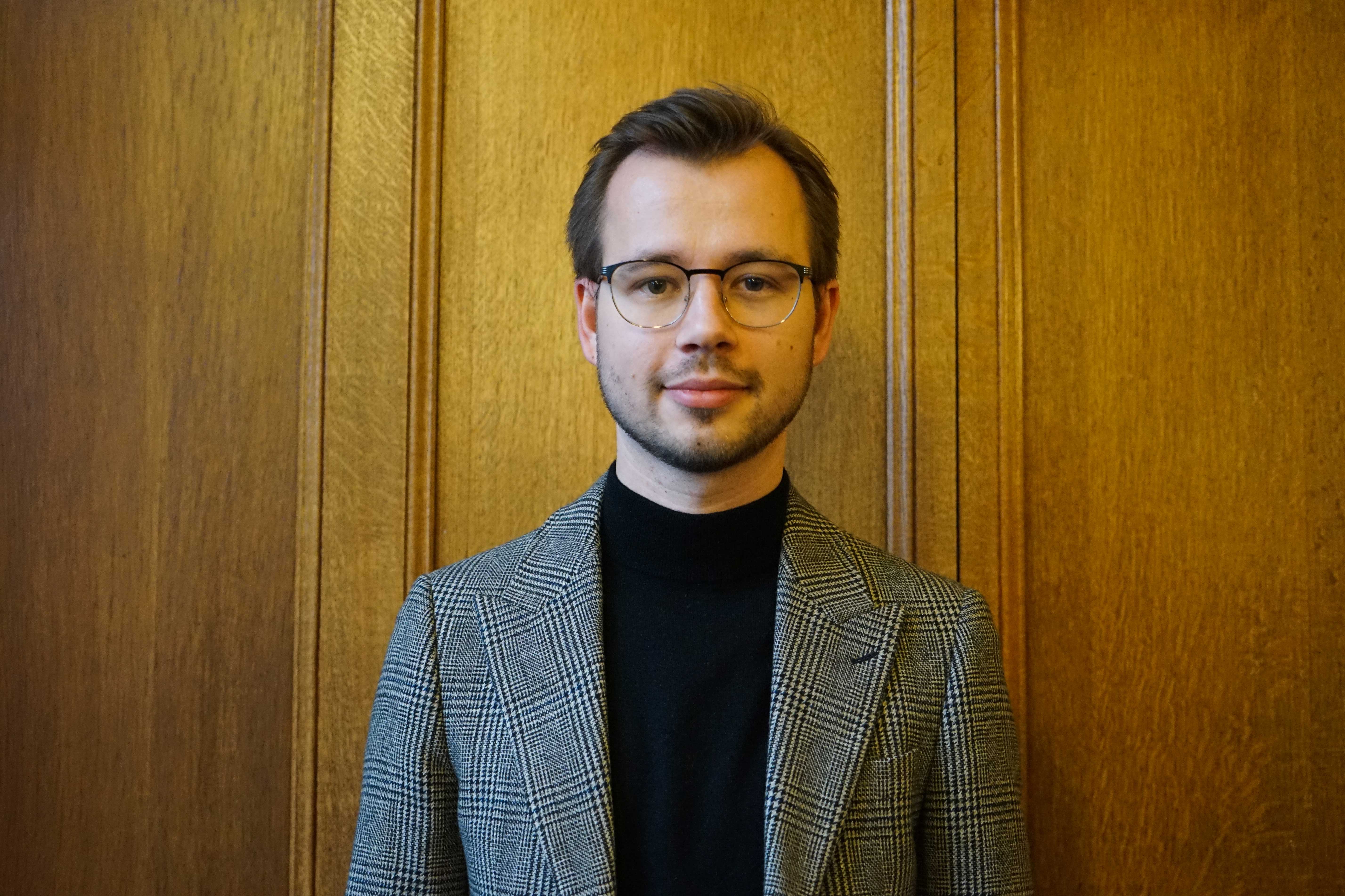 White male students in his twenties smiling tentatively at the camera, wearing a black and white houndstooth jacket with a black polo neck underneath. He has short dark brown hair, dark facial hair and glasses.