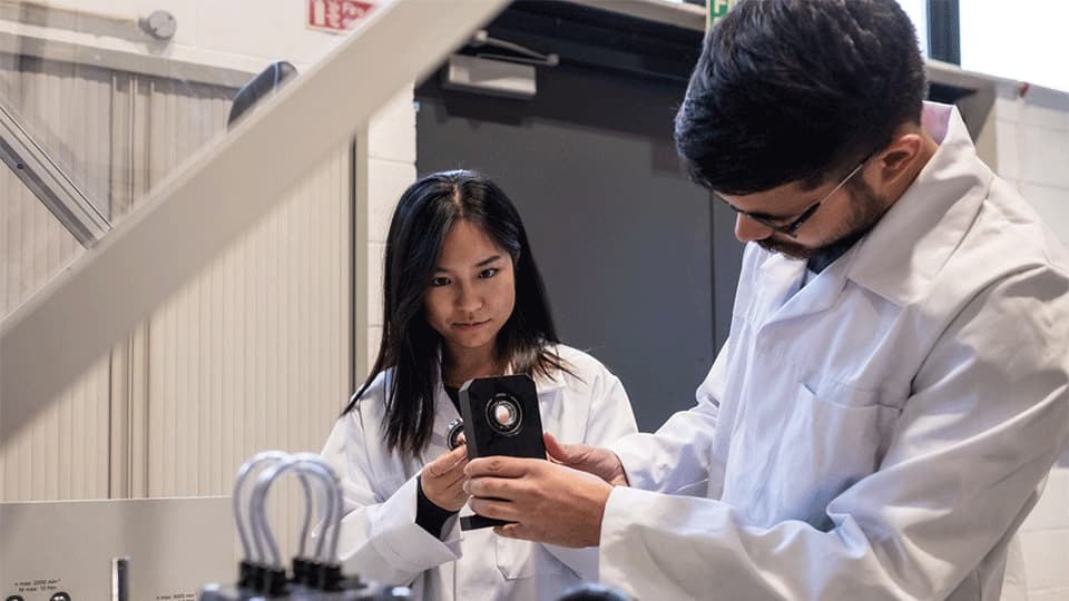 female and male student working in aerospace lab holding up equipment and wearing lab coats
