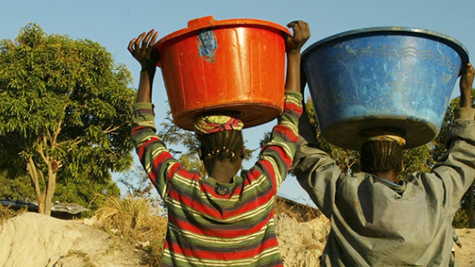 Two women of colour holding a red and blue bucket on their heads walking away from the camera