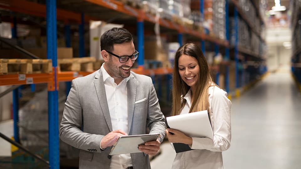 Two people in a warehouse looking at a tablet computer and a clipboard.