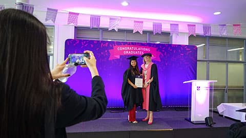 A graduate on stage holding a certificate from the Dean of Loughborough Business School. There is someone in the foreground taking a photo of them on a smartphone.