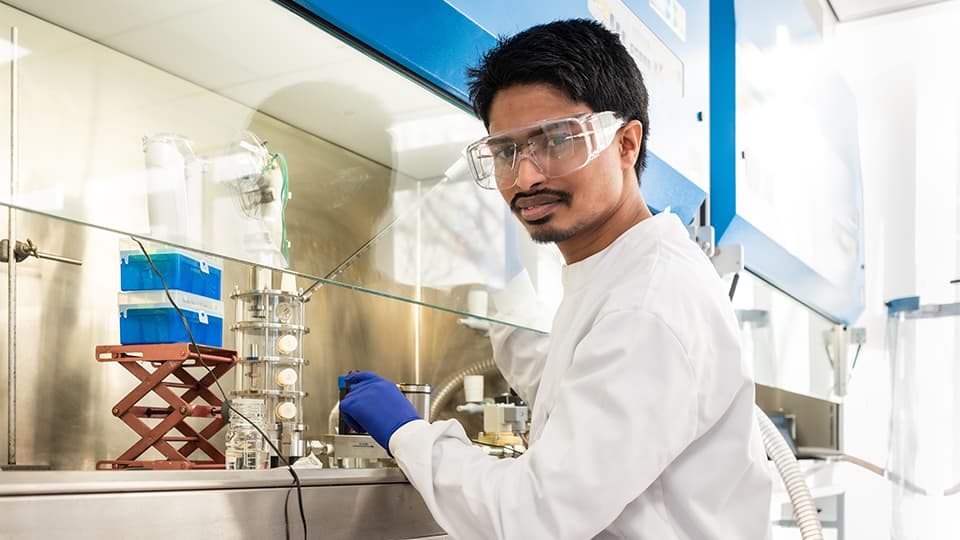 Student in lab coat, goggles and blue latex gloves looking at the camera and working in a lab