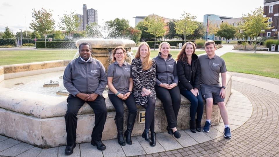 The friendly warden team at Loughborough are sat on the iconic foundation outside the Hazlerigg building smiling towards the camera and wearing university staff uniform,