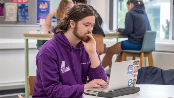 A male student wearing a purple hoody working on his laptop