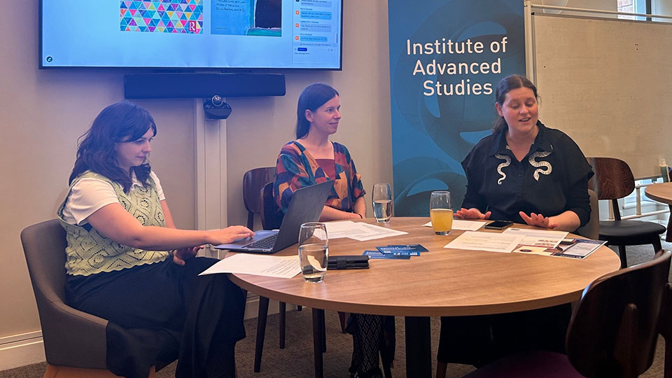 Three people sitting at a round table talking with a digital screen and pull-up banner in the background