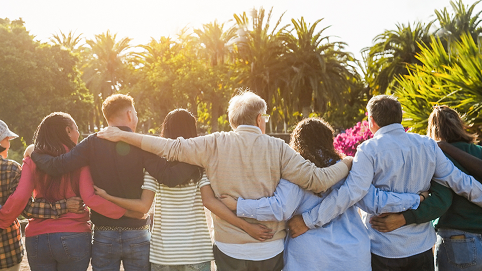 A group of people of different ages, races and genders with their backs to the camera with their arms wrapped around each other