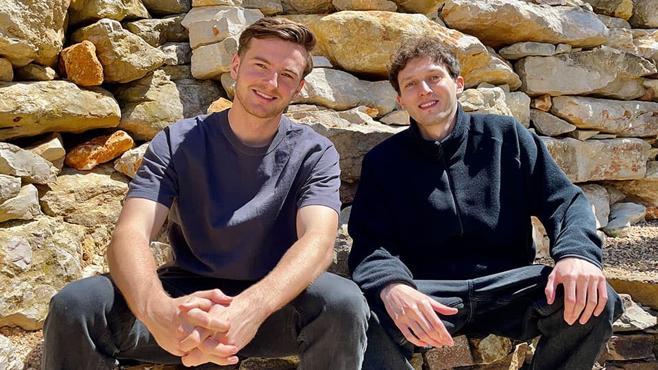 Ronan Harvey Kelly and Glenn Keller sitting next to each other smiling and they are sat on a wall with a wall behind them as the background.