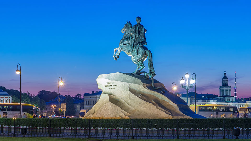 Statue of a man on a horse on top of a large stone, set against an evening sky with lampposts, buildings, and trees in the background.