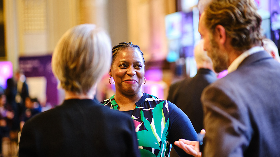 Photo of a guest listening to another colleague (with their back to the camera) speak whilst smiling