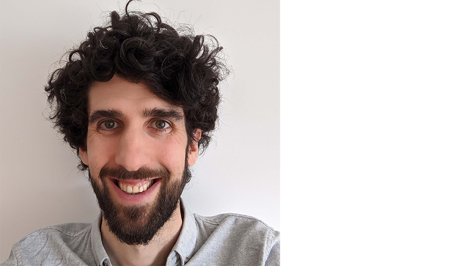 Close up headshot of Dr Anthony Kevins, smiling face on at the camera wearing a grey shirt