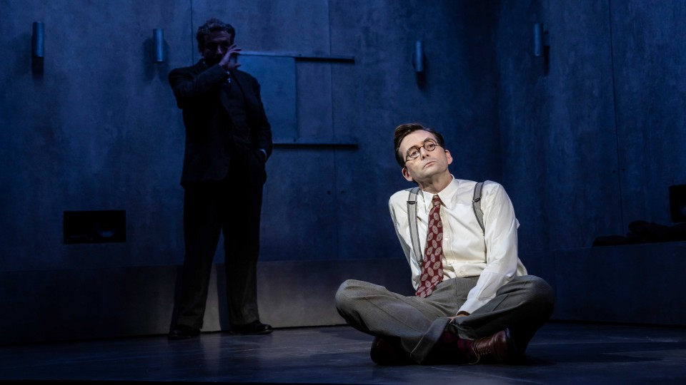 Photo of David Tennant sitting on a stage floor lit up by a spotlight, a man is standing behind him in shadow.