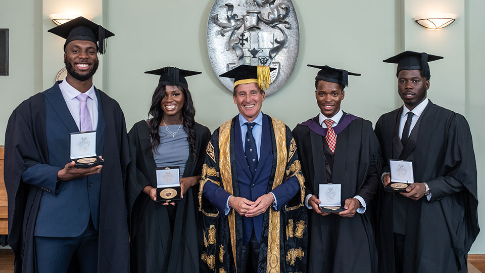 Four men and one woman stand in academic robes. They are holding medals