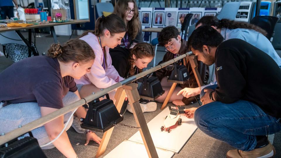 Photo of a group of girls crouched down on the floor looking at a toy car that is on a raised platform with lights above it