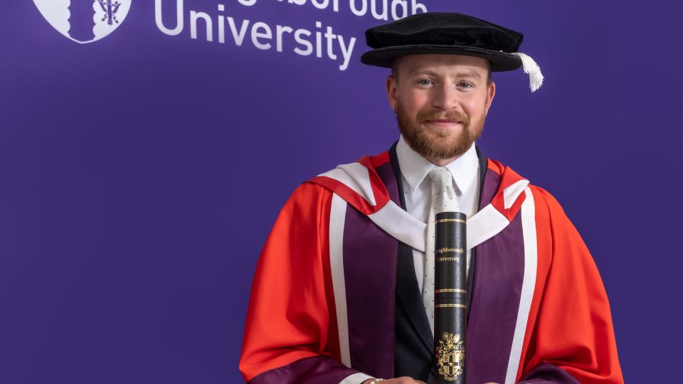 Adam dressed in graduation robes and cap stood smiling in front of a purple wall