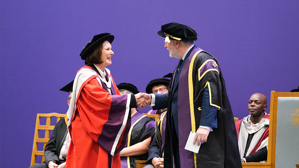 Professor Cooper shaking hands with the Vice-Chancellor on the graduation stage, both dressed in graduation hats and robes