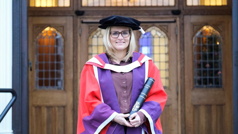 Sarina stood in front of the Hazlerigg Building front doors dressed in graduation robes