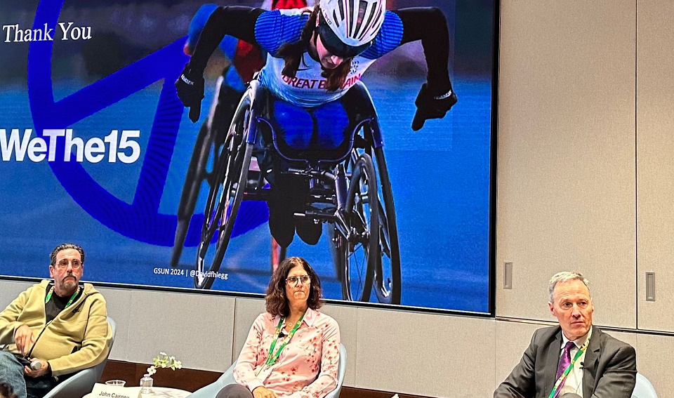 Professor Vicky Tolfrey is sat in front screen showing a wheelchair athlete