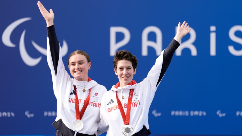 jenny holl and sophie unwin stood on a stage with their arms aloft with silver paralympic medals around their necks