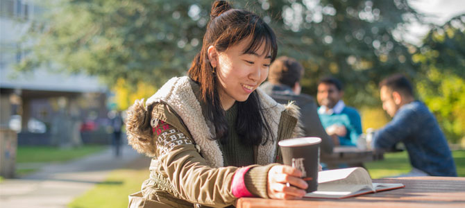 Smiling doctoral researcher sitting outside with a coffee, reading a book