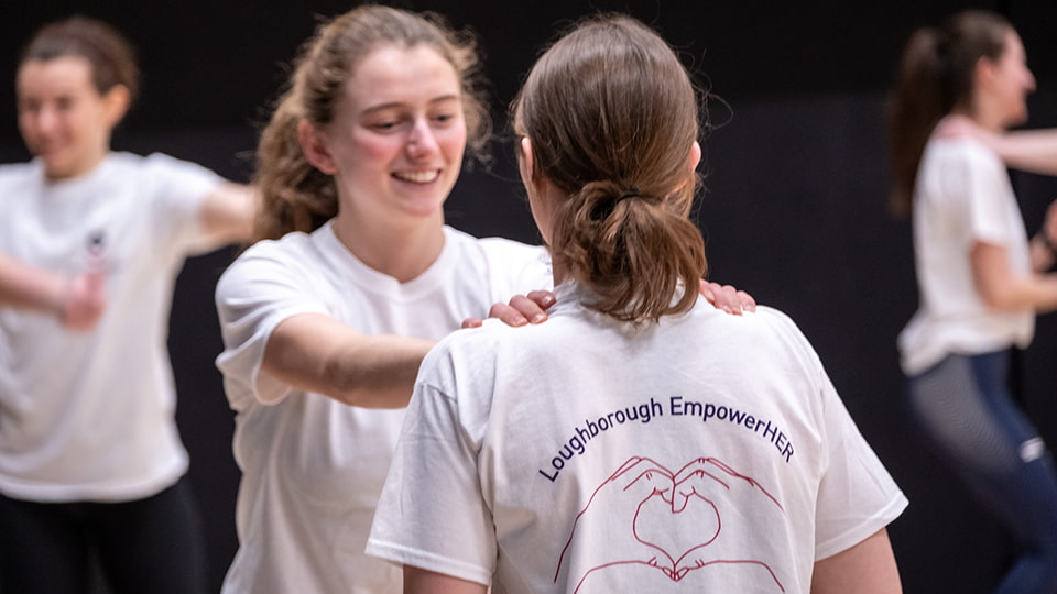 A woman smiling and holding her hands on another woman's shoulders who is wearing a Loughborough EmpowerHER t-shirt.