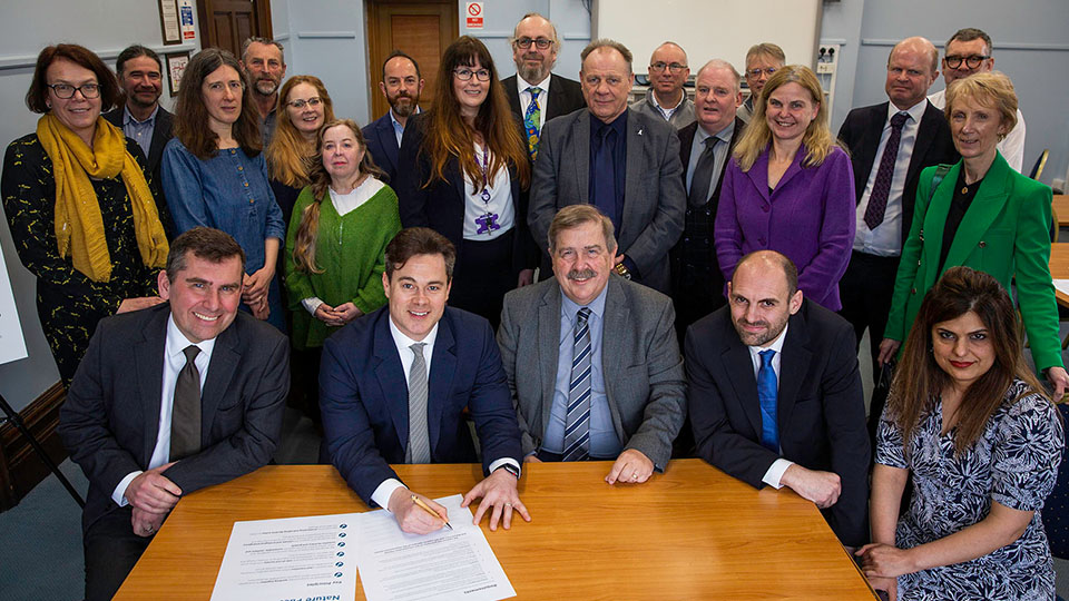 Photo of representatives from each of the signatories to the Pact sitting and standing around a table and smiling at the camera 